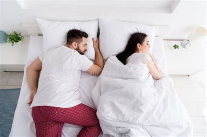 A couple in bed with the man sitting up texting while the woman looks suspicious in the background.