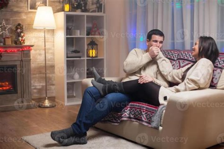 Husband sitting on couch with his wife and pleading for forgiveness with hands in prayer.